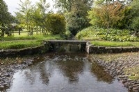 A ford, with cobbles, between a gravel drive. A footbridge runs beside the ford. Regency House, Devon NGS garden. Autumn