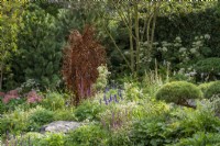Metal sculpture by Penny Hardy surrounded by planting of Veronica longifolia and Salvia 'Caradonna', Pinus sylvestris behind - Bodmin Jail: 60 Degrees East - A Garden between Continents, RHS Chelsea Flower Show 2021
