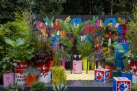 Planting in colourful containers surround seating area with black chairs and wall mural behind - Pop Street Garden, RHS Chelsea Flower Show 2021