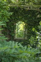 A framed mirror hung on an ivy clad fence to create the illusion of a window as a focal point at the end of a small garden. May.