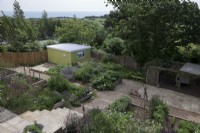 overview of the garden with sea view above yellow painted cabin. Herbaceous planting in raised beds with gravel pathways and paved stone step, with a mixture of flowers and vegetables . Cottage garden favourites including Digitalis purpurea - Foxglove,  Sweet Peas on   Hazel wigwam and woven hurdle fence and chicken coop.