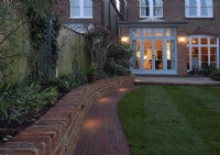 A London garden after a makeover with downlights on a brick wall,  a brick path around a raised bed and newly laid turf.