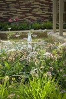 Herbaceous beds planted with perennials including  Pimpinella major 'Rosea' and Geum 'Petticoats Peach' next to a pond with fountains - The Stitcher's Garden, RHS Chelsea Flower Show 2022
