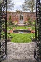 View through black metal gates into formal walled garden with gothic door, small round pool and containers of burgundy Tulipa