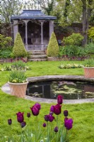 Ornamental summer house overlooking round pool in lawn lined with containers of burgundy Tulipa