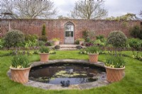 View into formal walled garden with gothic doorway with small circular pool lined with terracotta containers of burgundy Tulipa underplanted with yellow violas