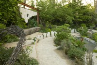 A sculpture made of nails, a small pond and seating area surrounded by trees and plants in the Samaritans' Listening Garden, a Show Garden designed by Darren Hawkes at the RHS Chelsea Flower Show 2023.