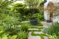 A circular metal water feature surrounded by ferns, trees and Yew hedge in the Myeloma UK - A Life Worth Living Garden designed by Chris Beardshaw at the RHS Chelsea Flower Show 2023