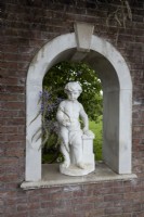 A stone statue of a child set within an archway in a brick wall with wisteria growing aorund it. Trago Mills show gardens, Devon, UK. May. Spring