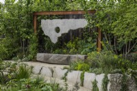 A wooden bench is backed by a sculpture made from salvaged materials, softened by perennials and hazel trees, Corylus avellana, to each side.