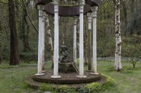 The Folly, a structure of columns with a tree stump in the centre and a statue of a cherub on top in a wooded garden. Marwood Hill Gardens. Spring. Devon. May. 