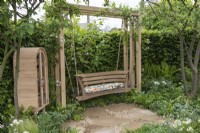 A quiet space with swingseat shaded by trees and edged in borders of ferns and white flowers. A wooden cabinet houses books, seeds and cushions.
