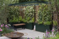 A formal courtyard is enclosed in willow and hazel fencing screens, supporting fragrant star jasmine, Trachelospermum jasminoides, which grows in tall planters with herbs. A water feature is crafted from reclaimed aluminium. Borders contain roses and herbaceous perennials.