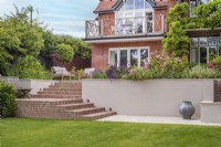View of brick steps and white rendered retaining wall leading up to raised terrace and house with mixed borders and furniture 