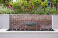 Verdigris basin fountain on brick wall of raised rendered water feature with terrace border with roses above.