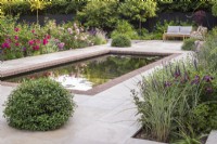 Brick edged rectangular pool overlooked by house on terrace of sandstone paving with garden furniture and border of David Austin Roses and Pittosporum topiary balls around pool