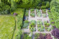 View over formal garden adjacent to meadow with gravel paths and rectangular beds containing Cynara cardunculus and Heuchera backed by hedges of Yew. June. Summer. Image taken with drone. 