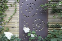 Metal screen with circular and spiral design attached to a contemporary wood boundary fence, white-flowered hydrangea in foreground