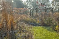 Sunlit rustic gazebo surrounded by ornamental grasses and perennials in winter.