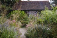 Paved path leads through small autumnal cottage style garden with perennials and grasses, and framed oak building behind.