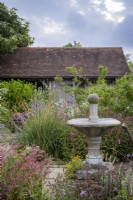 Small courtyard garden filled with late summer planting, with framed oak building behind and water feature, bird bath in foreground
