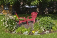 Red Adirondack chairs on wood plank deck bordered by Salvia - Silver Sage, Leucanthemum x superbum - Shasta Daisy 'Madonna', Hosta and Dahlia in backyard garden in summer.