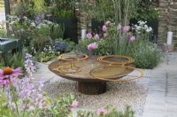 A weathered mild steel water feature with rings set in a gravel square surrounded by a mixture of planting influenced by aromatic and wildlife-friendly ornamentals in a design structure inspired by the parterre.  Rosa 'Princess Alexandra of Kent' in the background.  Landform Mental Wealth Garden - RHS Hampton Court Palace Garden Festival 2023. 