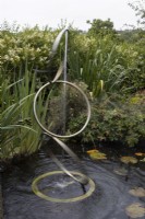 A rotating water feature, made of three interlinked hoops set in a pond. Water sprays downwards from the top hoop. A variety of water plants and other foliage is in the background. Harbour Lights, Devon NGS garden. July. 