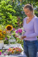 Woman arranging outdoor table with wreath and bouquets of flowers in vases. Flowers in bouquets are Hydrangea, sweet peas, Ammi majus and Nigella while wreath is made of Clematis seed heads, Dahlia and Rudbeckia.