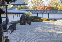 Area of raked gravel with placed stones. Surrounding walls are tiled. Single Japanese pine tree next to moss covered mounds. 