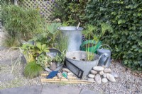 Metal basins, metal bin, watering can, bamboo sticks, pebbles, gloves, string, scissors, saw, Acorus, Juncus, Gunnera, Rodgersia, Nymphaea, Cyperus and Houttuynia laid out on the ground