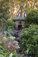 Prayer flags and a stone building with slate roof containing a water driven prayer wheel and waterfall over stones and rocks surrounded by planting of Pinus wallachiana, Rhododendrons, Persicaria amplexicaulis and Persicaria affinis 'Darjeeling Red' - The Trailfinders 50th Anniversary Garden September 2021