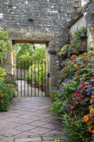A summer display of container-grown flowering and foliage plants including heliotrope, lantana and Fuchsia 'Thalia' line a paved path leading towards an arch and wrought iron gate at Bourton House Garden, Gloucestershire