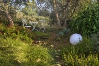 Mediterranean garden view with mass planting of drought tolerant plants, bushes and trees. On foreground is a Pennisetum villosum grass, decorative elements as wooden stairs, white ball and romantic setting with table and chairs. 

Italy, Tuscan Maremma, Orbetello
Autumn season, October
