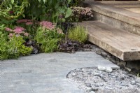 A clay brick paved path leading to wooden timber steps - planting in the border of Sedum spectabile 'Brilliant',  Sedum 'Mr Goodbud', Sedum telephium 'Matrona' and Ajuga reptans 'Black Scallop'  