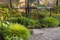 An old industrial urban wasteland redesigned into a new urban garden with reclaimed cobble and stone paving slabs and gravel for rainwater drainage - mixed perennial planting with Hakonechloa macra and reclaimed old pipes sculpture and a brick wall 