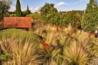 Mediterranean garden view at the entrance zone decorated with violin sculptures, grassland with Stipa tenuissima and Olive tree in the iron container in autumn time, sunlit with warm evening light.

Italy, Tuscan Maremma, Orbetello
Autumn season, October
