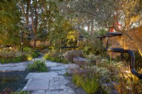 An old industrial urban wasteland redesigned into a new urban garden lit up at night with reclaimed cobble stone paving - pond - mixed perennial planting