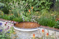 Copper metal rill on top of a low stone retaining wall flowing into a small circular bird bath pool -  mixed perennial planting of Echinacea, Kniphofia 'Papaya Popsicle', Helenium, Achillea, Erigeron
