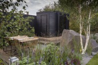 Small courtyard garden with a reclaimed wood floor patio area - table and chairs - a timber wooden deck with a black painted wood sauna cabin - living roof planting of wildflowers and herbs