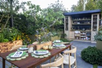 An outdoor kitchen with a paved patio area with dining table. A Ficus carica - fig tree and charred wood wooden raised beds with mixed perennial edible planting - 
