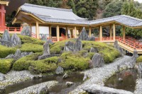 View across paved area with small pools and placed rocks with bed of Rhododendron (not in flower). Steps and pavillion of temple building. 
