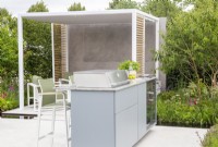 A modern contemporary outdoor kitchen area on a white stone paved patio with a covered pergola
