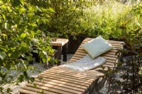 A wooden recliner with cushion and blanket and a small table in a woodland garden mixed perennial planting and ornamental grasses
