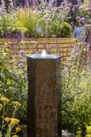 A stone column water feature with mixed perennial planting including Achillea millefolium, Verbena bonariensis and purple flowered Salvias