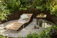 An enclosed relaxing area with gravel surface and wooden raised beds,  wooden recliner with blanket and cushion in a woodland garden with mixed perennial planting 