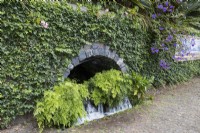 An arching cave with waterfall beneath, set within an ivy covered wall with ferns growing beneath. A cobbled path is in front. Monte Palace Gardens, Madeira