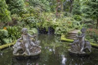 Two figurative statues in a pond, with small waterfalls in the background, steps to the right and lush tropical planting in the background. Monte Palace Gardens, Madeira
