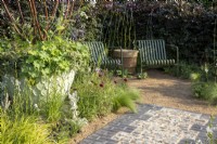 A stone flint and grey brick path leading to a gravel surface seating area, container with Multi-stemmed Prunus serrula and Alchemilla mollis and mixed perennial planting border including Cosmos atrosanguineus