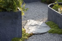 A gravel surface with a rock in between galvanised metal container and raised pond 